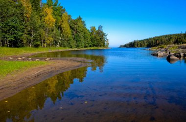 Mavi, şeffaf, büyük taşları olan sığ bir nehir. Mavi açık gökyüzü. Kıyılar boyunca yeşil çam ormanı ve sarı huş ağaçları vardır. Sol kumlu kıyı sakin suya yansıyor..