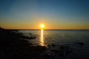 Gölün üzerinde parlak gün batımı. Mavi gökyüzündeki sarı güneş. Karanlık suda güneş yolu. Kaya kıyısı.