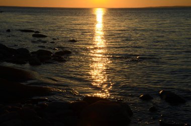 Gölün üzerinde turuncu gün batımı. Sakin suda güneşli bir yol. Karanlık sularda siyah taşlar.