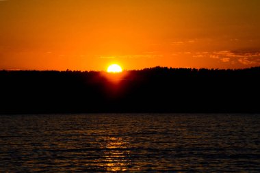 Gölün üzerinde parlak gün batımı. Turuncu güneş ormanın üzerinde batar. Sarı günbatımı. koyu su.