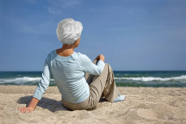 Ηλικιωμένη γυναίκα που κάθεται στην άμμο στην παραλία και βλέμματα στην απόσταση στη θάλασσα ορίζοντα — Φωτογραφία Αρχείου