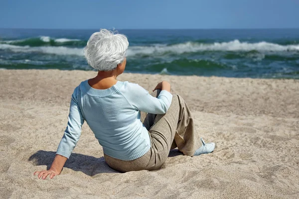 Ηλικιωμένη γυναίκα που κάθεται στην άμμο στην παραλία και βλέμματα στην απόσταση στη θάλασσα ορίζοντα — Φωτογραφία Αρχείου