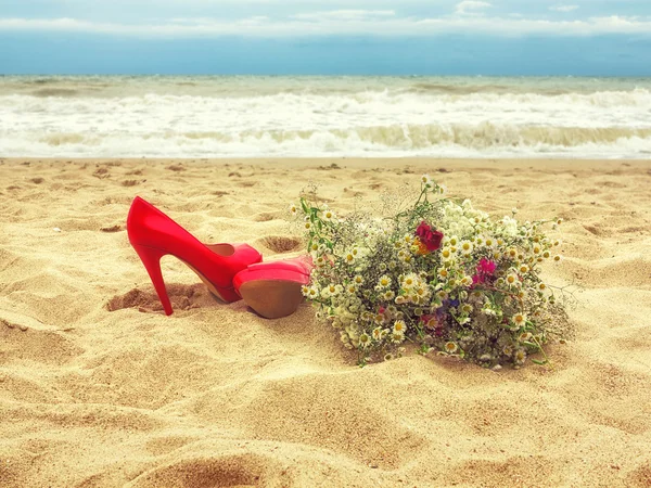 Red shoes and a bouquet of wild flowers on the sand near the sea — Stock Photo, Image