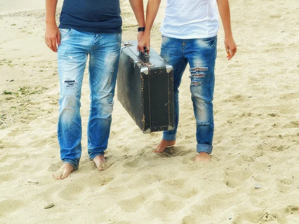 Male and female feet on the sand near the sea with a leather suitcase, image in the sunny  trendy vintage style — Stock Photo, Image