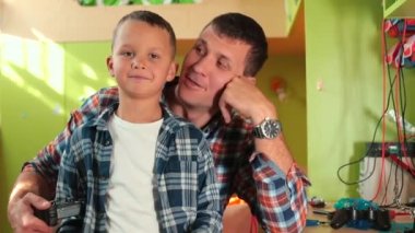 Baba ve oğul evde poz veriyorlar. Baba ilgiyi gösterir, yanağa dokunur.