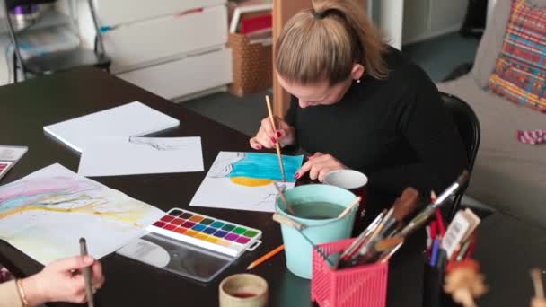 De student tekent een tekening aan tafel. Ze borstelde verf op papier borstel — Stockvideo