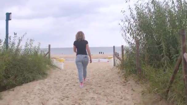 Ragazza torna si muove lungo la strada per la spiaggia. Prende passi sulla sabbia verso il mare. — Video Stock