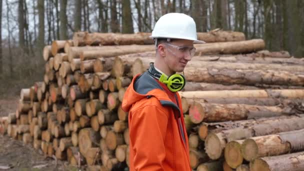 Um trabalhador madeireiro em um chapéu duro posa perto dos troncos das árvores. Pausa o trabalho. — Vídeo de Stock