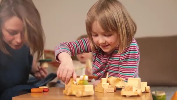 Glada lilla flicka spelar leksaker på bordet. Mot bakgrund mamma med barn. — Stockvideo