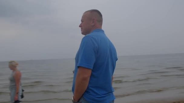 Человек стоит на берегу моря в облачный день. Синяя футболка. Он смотрит вперед, мечтает. — стоковое видео