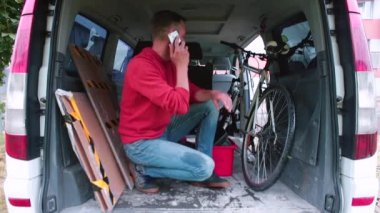 Teslimatçı, arabanın bagajında telefonla konuşuyor. İletişim kurmadan önce maske takıyor.