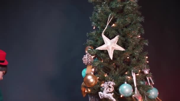 El adolescente se levanta cerca del árbol, cuelga un juguete redondo en el árbol. — Vídeo de stock