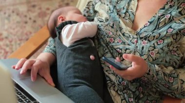 Yeni doğmuş bebeği ve telefonu olan bir kadın evinde dizüstü bilgisayarı olan bir sandalyede oturuyor..