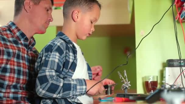 De actieve zoon soldeert de delen van het elektrische apparaat aan de tafel. — Stockvideo