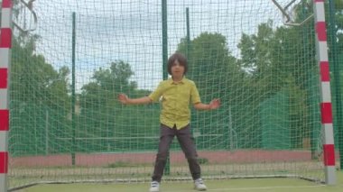 Uzun saçlı ergen, futbol kalesini savunuyor. Patlamaya hazırlanıyor. Vücut hareketleri yapıyor.