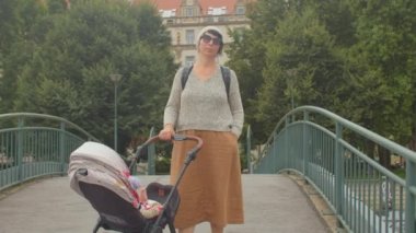 Elbiseli genç anne, elleri köprüde bebek arabasını tutuyor. Gözlükler takılı..