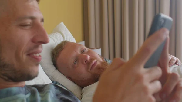 Доброе утро в спальне. Счастливый молодой гей со своим любовником пользуется телефоном. — стоковое фото
