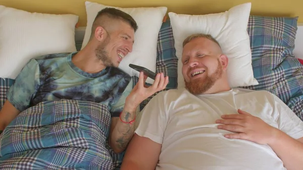 Homo dating app. Twee bebaarde mannen liggen in bed met een telefoon, zoenen. — Stockfoto
