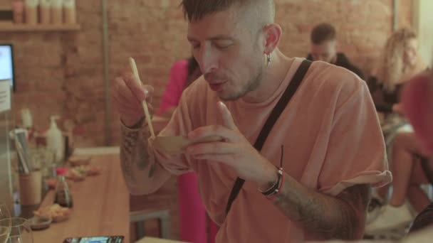 Голодний молодий чоловік швидко їсть частину азіатської їжі в міському кафе.. — стокове відео