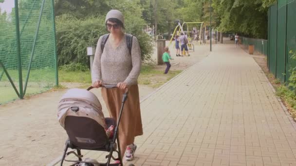 Mujer gafas conduce cochecito a lo largo de la acera en el patio de la madre muchos niños — Vídeos de Stock