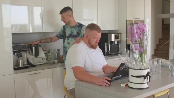 Two gay men kitchen man beard uses computer table partner makes smoothie — Stock Video