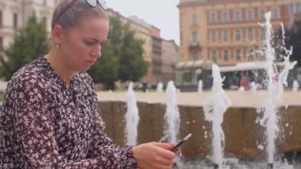 Flickan är ängsligt läsa nyheterna med hjälp av telefonen vid stadens fontän. — Stockvideo
