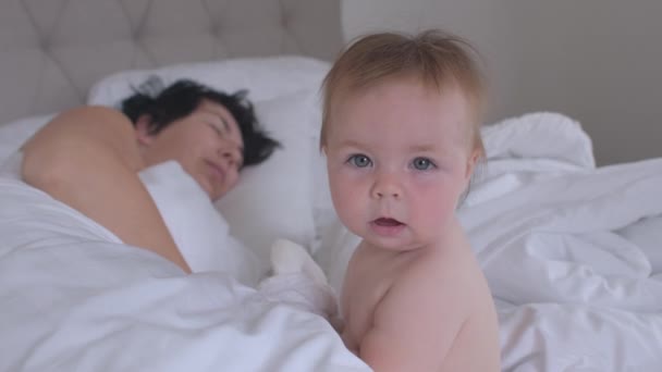 Leuke baby poseren met slapende moeder in een wit bed. Een droom delen met een kind — Stockvideo