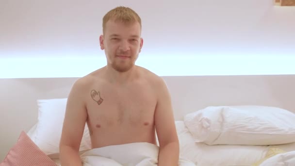 A young man uses a movie clapperboard while sitting in bed with a naked torso. — Stock Video