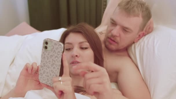 De cerca. Pareja joven enamorada usa el teléfono mientras está acostada en la cama. Aplicación de citas. — Vídeos de Stock