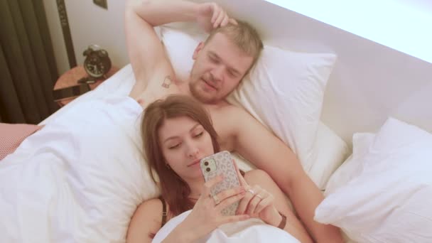 Νεαρό ερωτευμένο ζευγάρι χρησιμοποιεί το τηλέφωνο ενώ είναι ξαπλωμένο στο κρεβάτι. Εφαρμογή ημερομηνίας. — Αρχείο Βίντεο