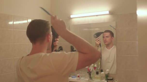 Facet używa suszarki do włosów do stylizacji łazienki. Patrzy w lustro, czesze włosy. — Wideo stockowe