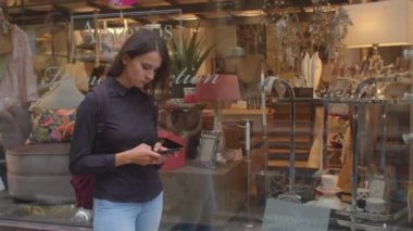 Kız dükkan penceresinin yanındaki telefonu kullanıyor. Ekrana bakıyor, düğmelere basıyor..