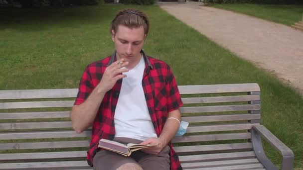 Lonely young man smokes a cigarette and reading a book. Outdoors lifestyle. — Stockvideo