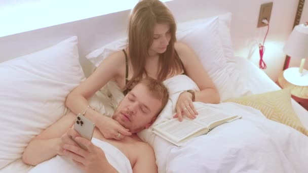 El hombre usa el teléfono en la cama en sus manos. Ella lee el libro, lo abraza con su mano — Vídeos de Stock