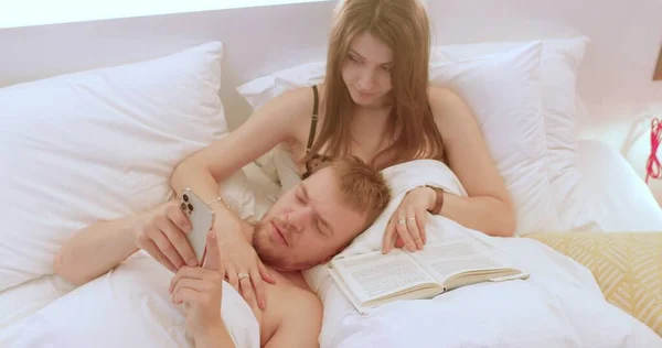 Man uses phone in bed in his hands. She reads book, hugs him with her hand — Fotografie, imagine de stoc
