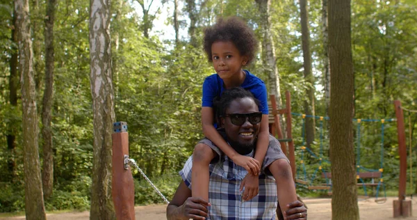 Papa avec mignon fils bouclé sur les épaules parle à la caméra sur le terrain de jeu. — Photo