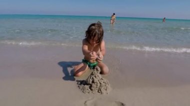 Çocuk sahilde kumdan kale inşa ediyor, insanlar suda dinleniyor.