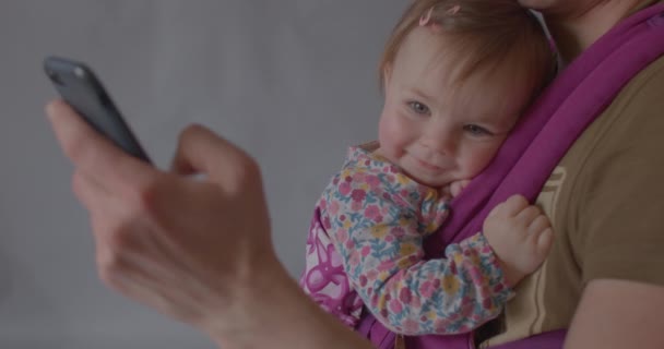 Linda niña sonríe coqueta mientras está sentada en un portabebés ergonómico. — Vídeos de Stock