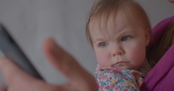 O retrato do bebê bonito cuidadosamente olha para o fechamento do telefone. Encontrar aplicativo móvel babá. — Vídeo de Stock