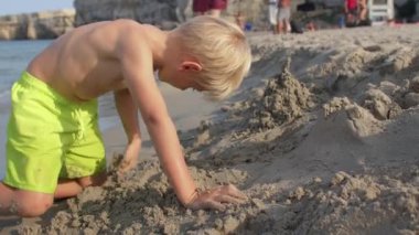 Bir çocuk deniz sahilinde çıplak elleriyle kumu kazıyor..