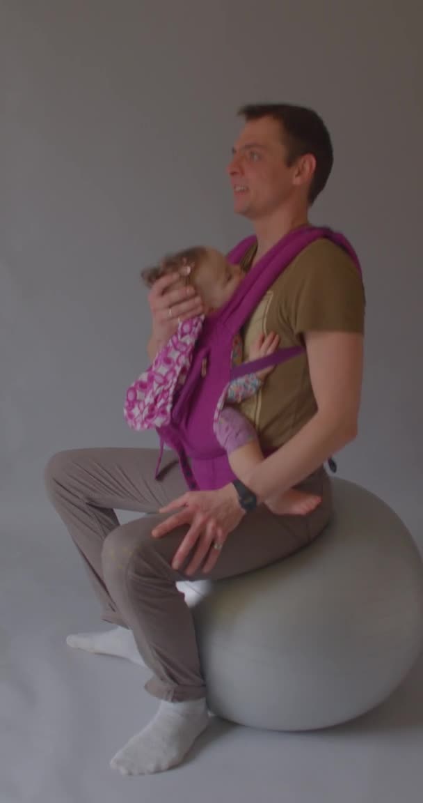 Père fait basculer l'enfant sur fitball. Porte-bébé en tissu doux. Vidéo verticale. — Video