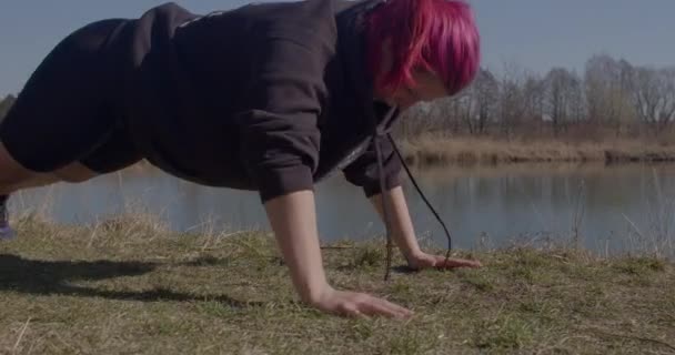 Una donna attiva che fa flessioni da terra sul fiume. Tiene il corpo, piega il gomito delle braccia. — Video Stock