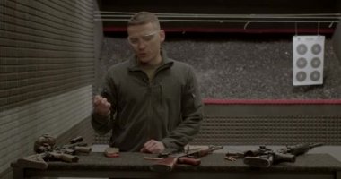 Erkek eğitmen elinde fişek tutuyor, askeri silahlardan bahsediyor.