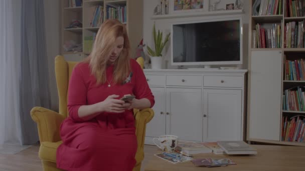 Κοκκινομάλλα νεαρή γυναίκα με ένα τηλέφωνο στα χέρια της πίνει τσάι σε μια κίτρινη πολυθρόνα — Αρχείο Βίντεο