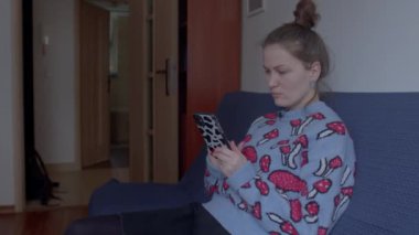 Bir kadın odanın kanepesinde otururken telefonu kullanıyor. Ekrana bakıyor, düğmelere basıyor. Saçlar at kuyruğu şeklinde yapılır. Taşınabilir internet kullanımı.