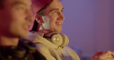 Bir arkadaşıyla konuşurken boynunda kulaklık olan bir adamın portre yüzü. Bokeh 'deki adama bakıyor. Elinde bir oyun kumandası var. Yüze hareket ışığı, aksiyon oyun odası.