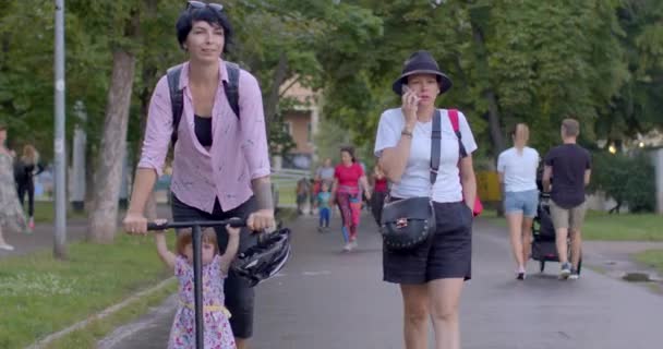 娘と一緒にスクーターで母親の動き 彼女は彼女の友人と歩道に沿ってロール 彼女は電話をかけ 一歩を踏み出す 都市公園を通って移動する プラハ ストロモフカ — ストック動画