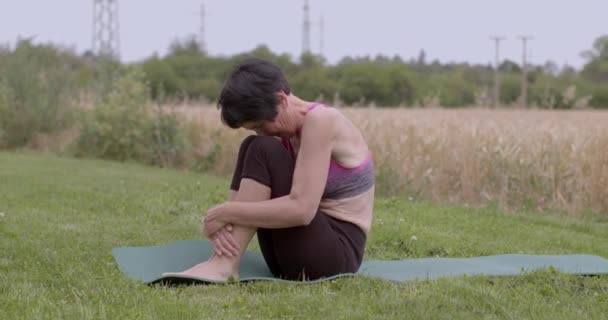 女性のシニアは訓練で彼女の上の足を上げる 彼女は体重体で背中に落ち 持ち上げます リハビリテーションにおけるボディトーンの増加 屋外で運動をする — ストック動画