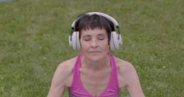 Mutlu sporcu kadın. 60 yo müzik rahatlığıyla meditasyon yapıyor. Dışarıdaki insanların sağlıklı yaşam tarzları. Yaşlı insanlar için meditasyonun faydaları. Kanserden sonraki yaşam. 