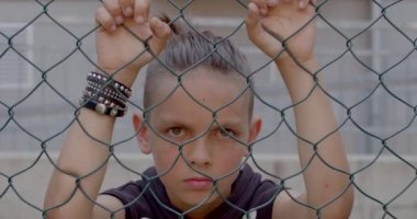 Kızgın çocuk ellerini çitin duvarında tutuyor. Çitlerle çevrili alandaki kafeslere bakıyor. Serbest bırakılmayı bekliyor. Elindeki perçin kayışı..